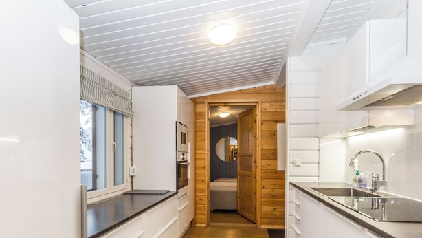 Une cuisine étroite et moderne, avec des meubles blancs, une porte en bois donnant sur une salle de bain et un tapis gris sur le sol.