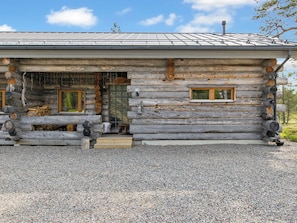 Une cabane en rondins avec une allée de gravier, un banc en bois à l’extérieur et une fenêtre donnant sur la forêt.