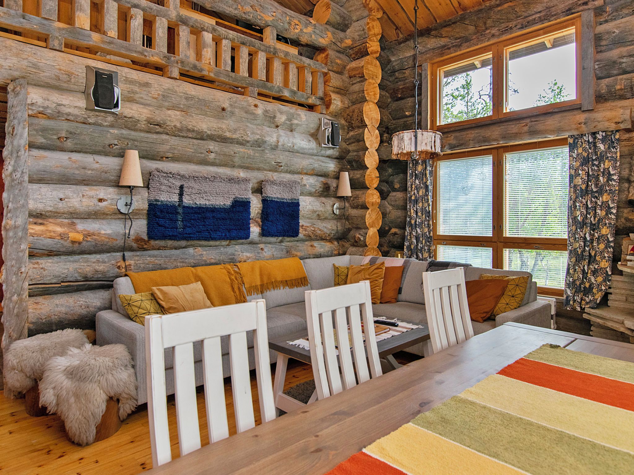 A log cabin interior with a sofa set, a wooden dining table, and a large window with curtains.