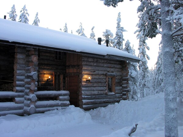 A log cabin with a snow-covered roof, wooden walls, and a small porch area with two illuminated windows.