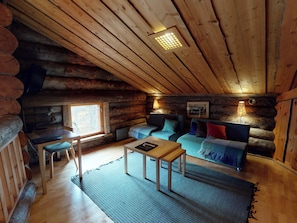 A log cabin interior with a wooden ceiling, a sofa, a table, and a window.