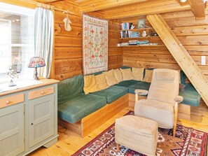 A cozy cabin interior with a green sofa, a wooden chair, a small table with a lamp, and a bookshelf.
