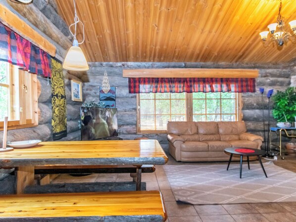 A log cabin interior with a wooden dining table, a sofa, and a window with a plaid valance.