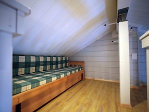 Ein kleiner Raum mit schrägen Wänden, einem hölzernen Bett und einer weißen Wand.