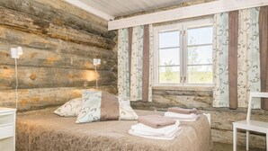 Ein Schlafzimmer in einer Blockhütte mit einem Bett, Kissen, einem Sessel und einem Fenster mit Vorhängen.