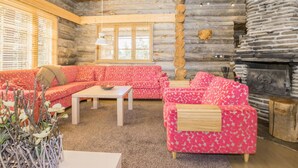 Ein Interieur einer Blockhütte mit Holzbalkendecke, einem Steinkamin, einem pinkfarbenen Möbelstück mit Muster und einem Holztisch.