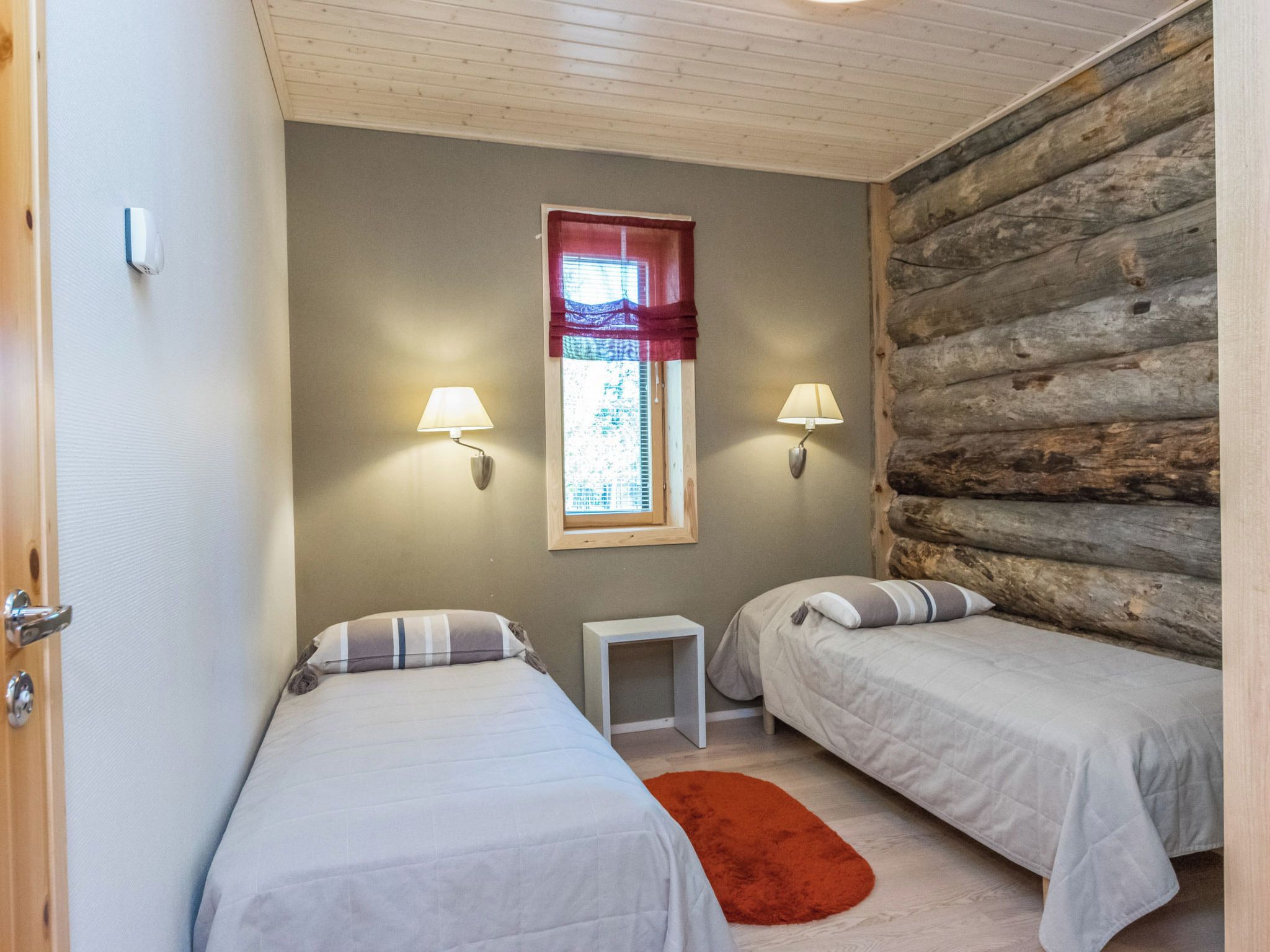 Une chambre avec deux lits, une petite table, une fenêtre avec un rideau et un mur en bois.