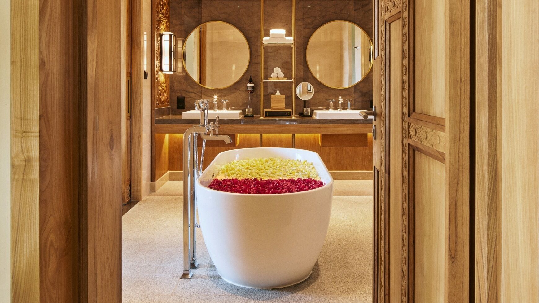 Un baño lujoso con una bañera blanca grande llena de pétalos de rosa, un tocador de madera con dos lavamanos y dos espejos grandes encima.