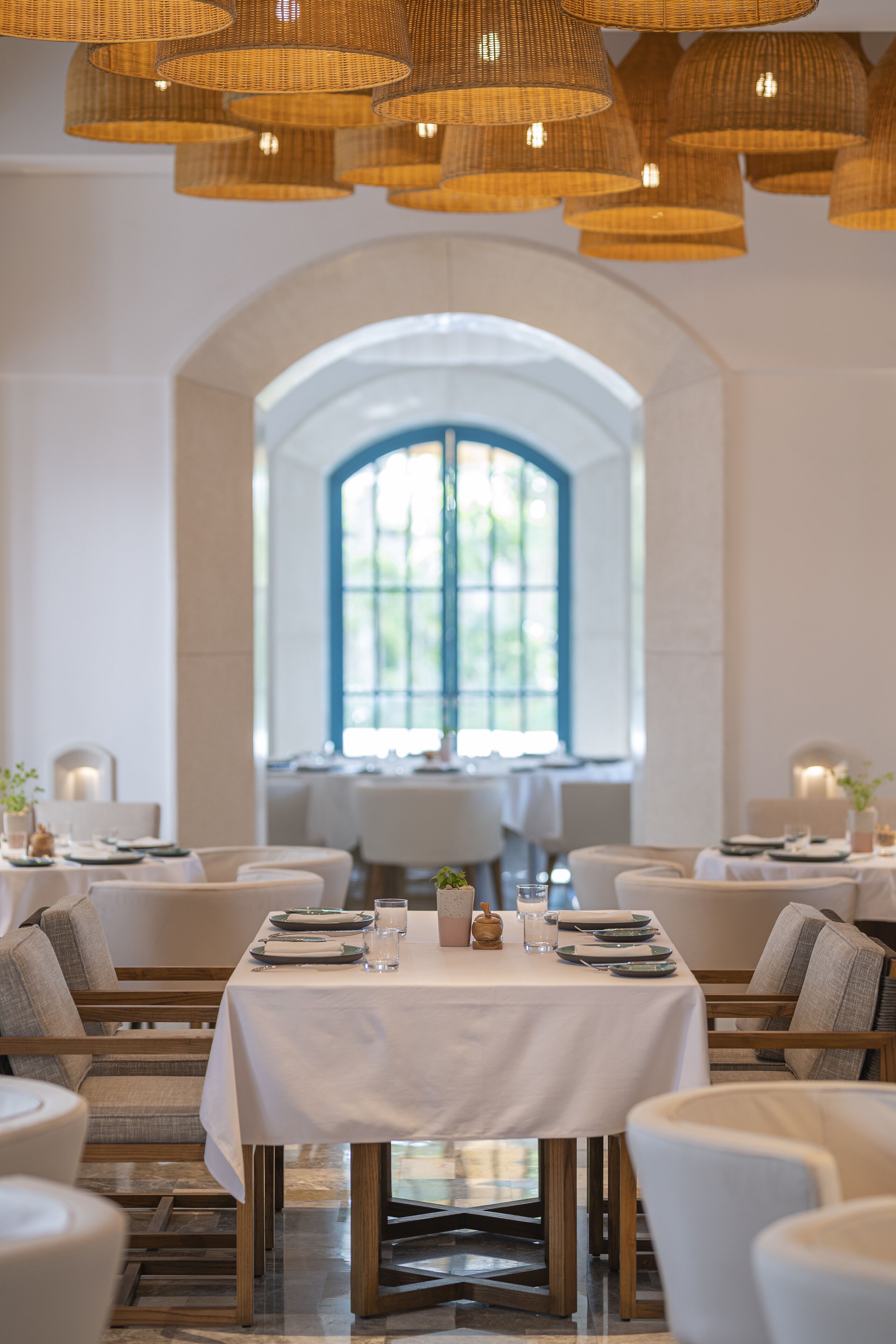 A dining area with a white table set for two, surrounded by chairs, and a large arched window in the background.
