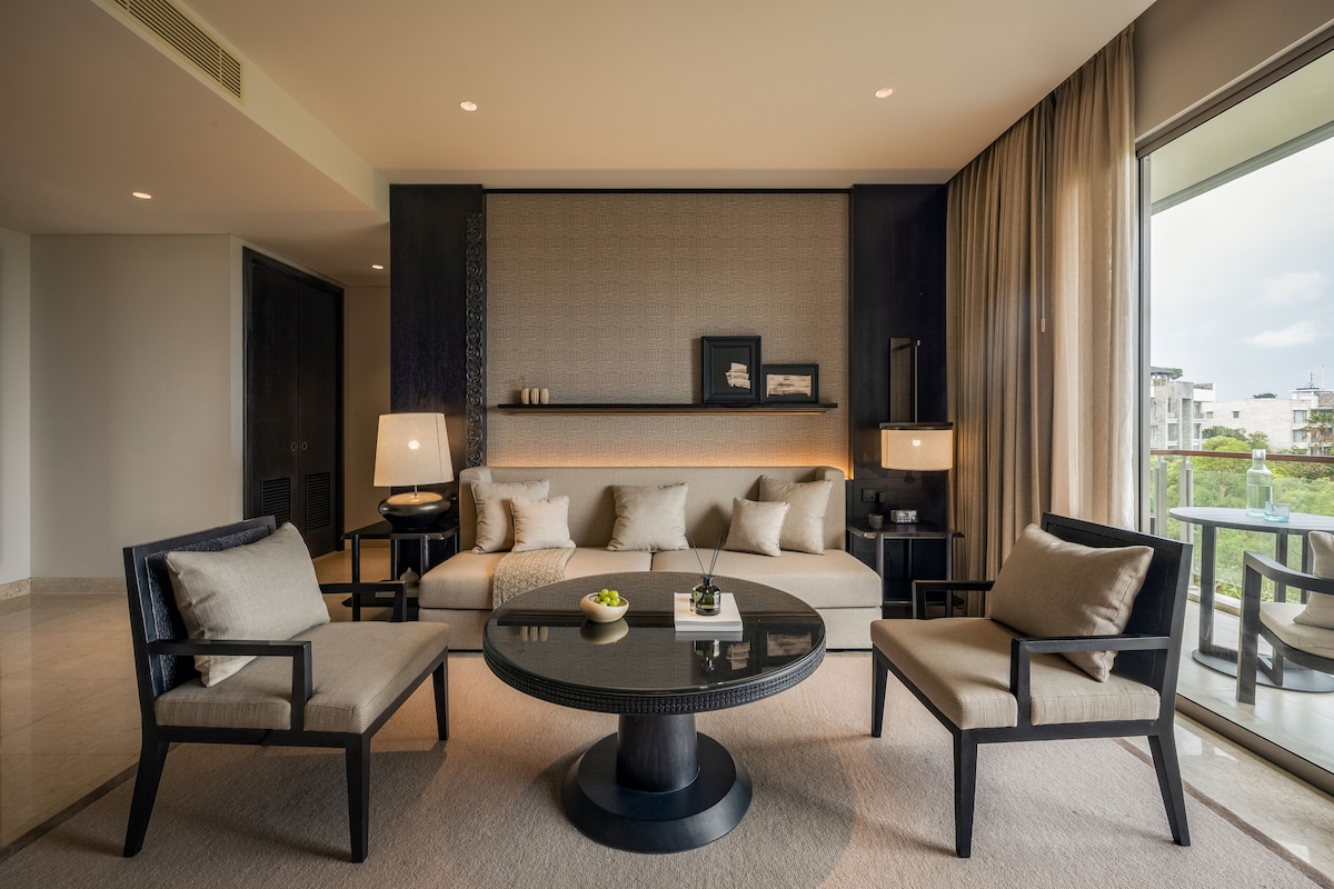 A modern hotel room with a large bed, two armchairs, a round coffee table, and a view of the city through large windows.
