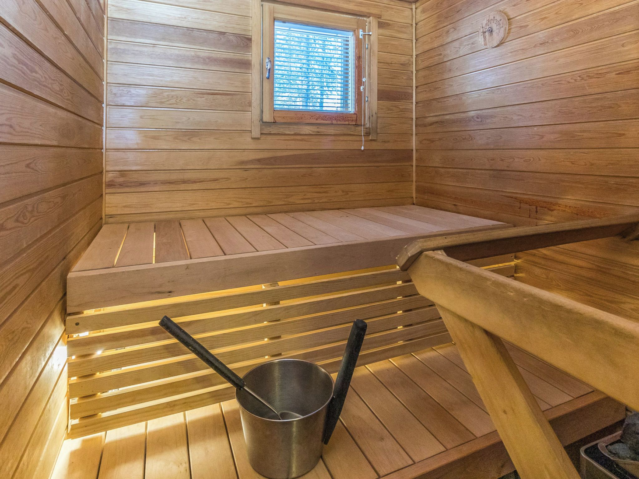 Een sauna met houten wanden, een bank, een raam met zonwering en een metalen emmer.