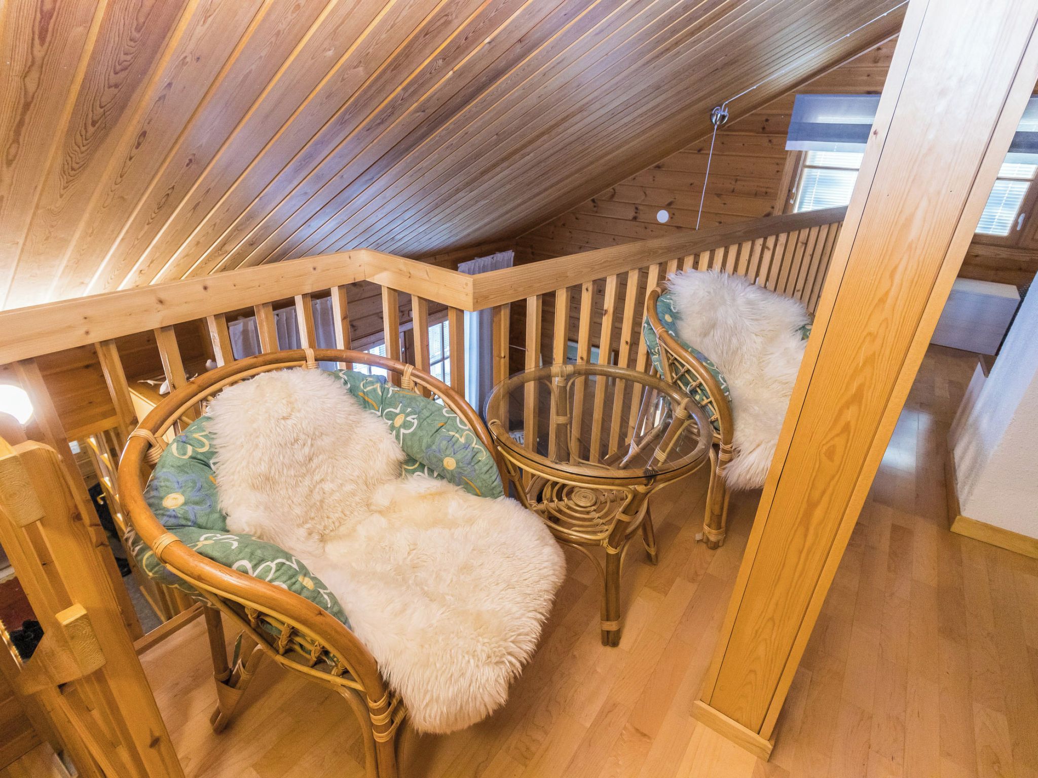 Een houten interieur met twee rieten stoelen en een met kussens uitgeruste bank, een houten leuning en een schuine balkenplafond.