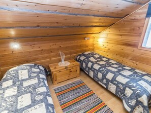 A small, cozy room with wooden walls and a sloped ceiling. There are two beds with patterned bedding, a small wooden nightstand, and a colorful striped rug. - Aihki 1 by Interhome (Inari)