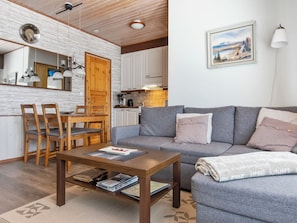Una sala de estar acogedora con un sofá gris, una mesa de comedor de madera y sillas. Se ve una cocina al fondo con armarios blancos y una puerta de madera.