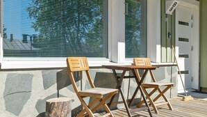 Eine Holzterrasse mit zwei Stühlen und einem Tisch, ein Fenster mit Jalousien und eine Tür mit einem Schild.