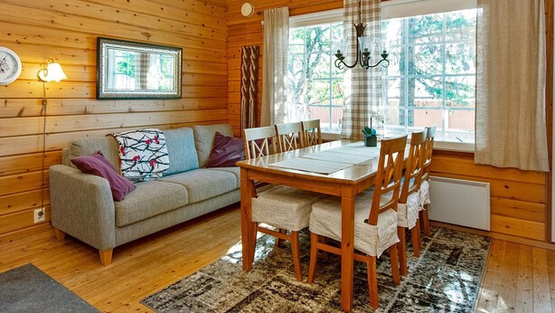 Interior acogedor de una cabaña con una mesa de comedor de madera, sillas, un sofá y un ventanal que ofrece vistas a la vegetación.