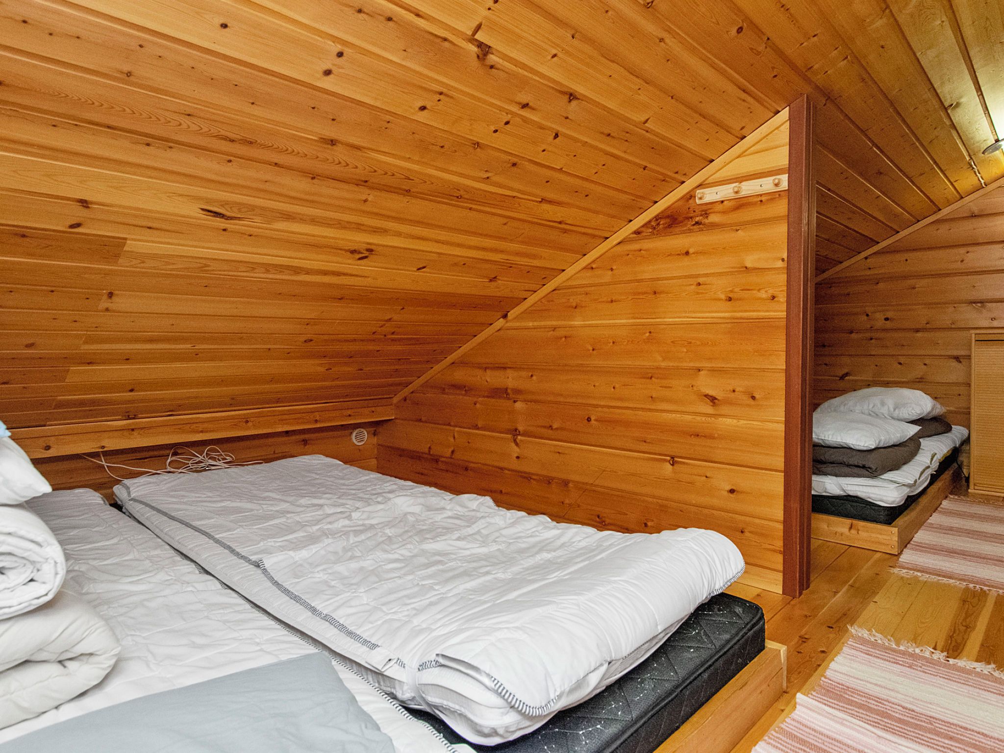 A wooden cabin bedroom with a single bed, a wooden ceiling, and a small storage area with folded bedding.
