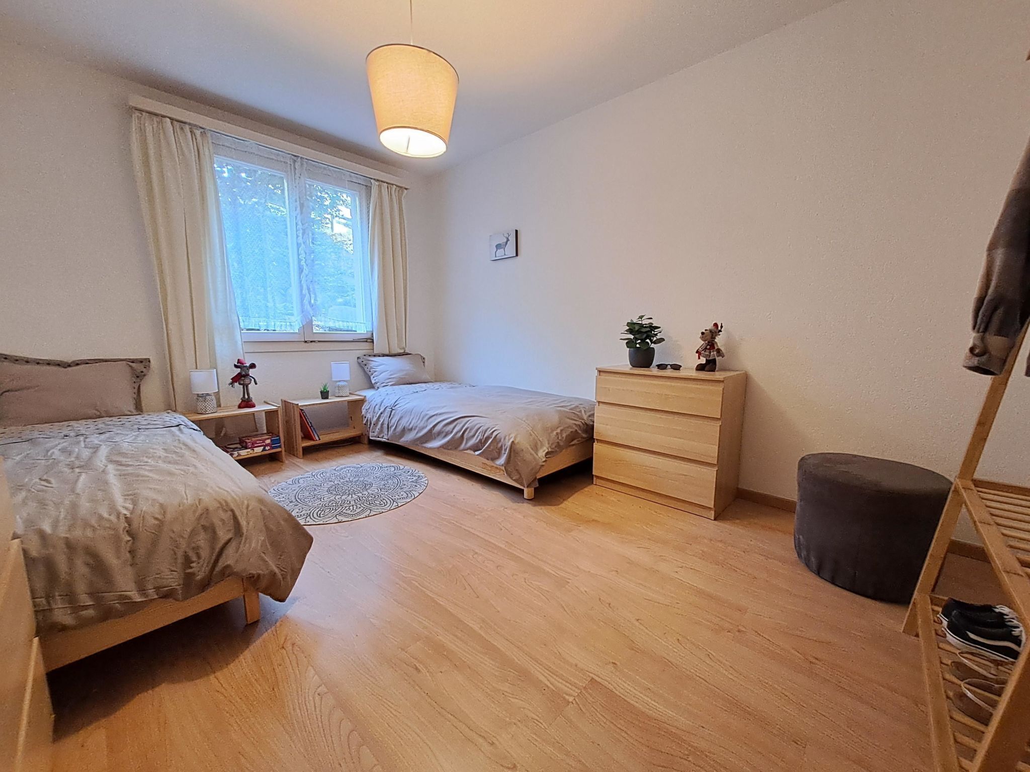 Un dormitorio con dos camas, un aparador, una ventana con cortinas, una alfombra pequeña y una estantería con objetos.