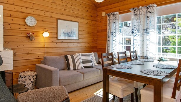Interior acogedor de una cabaña con paredes de madera, un sofá, una mesa de comedor y una ventana con cortinas.
