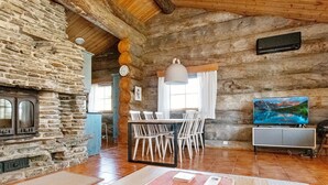 Un interior rústico con chimenea de piedra, vigas de madera en el techo, una zona de comedor con sillas blancas y un televisor de pantalla plana montado en la pared.
