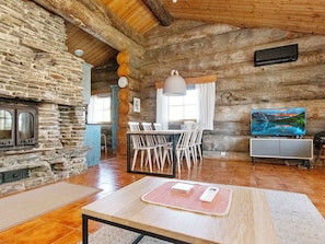 Un interior rústic amb una paret de pedra, un paviment de fusta, una llar de foc, una zona de menjador amb cadires, una televisió i una finestra gran.