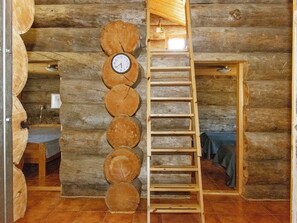 Interior de una cabaña de troncos con una escalera de madera y un reloj fijado en una pared de tronco.