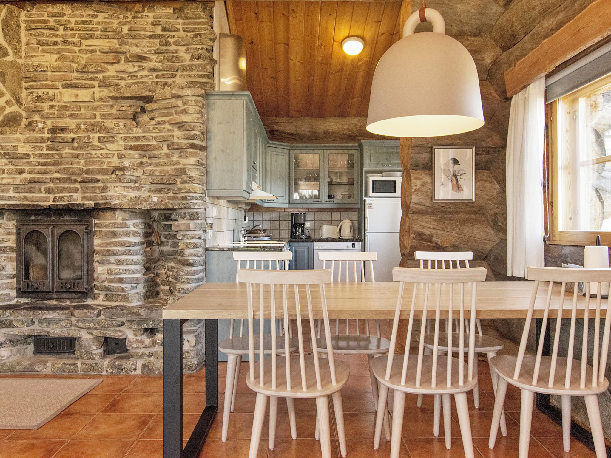 A rustic kitchen with stone walls, wooden ceiling, and a dining area with white chairs and a table.