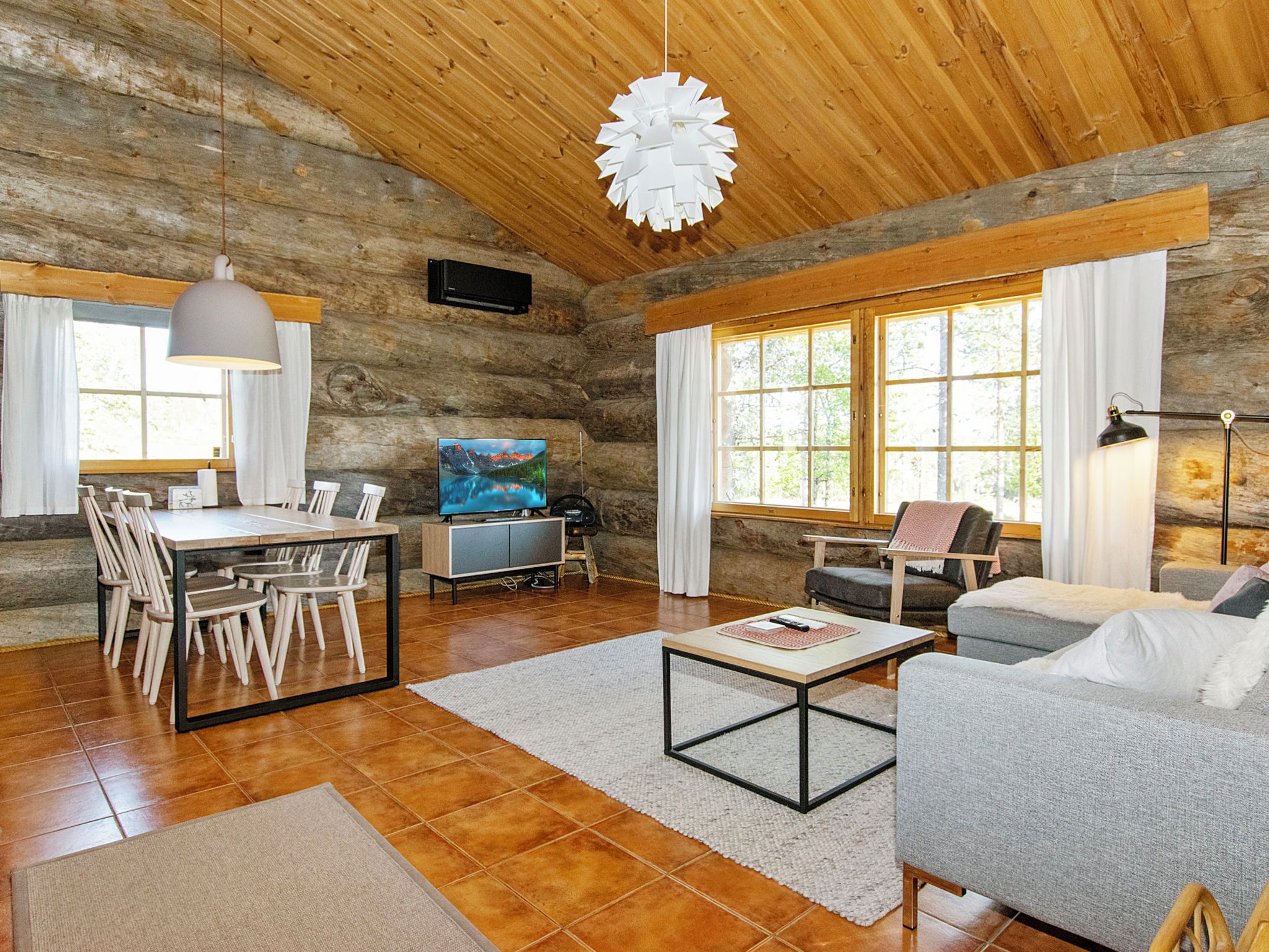 A rustic cabin interior with a dining area, a living room with a sofa and armchairs, and a large window with curtains.