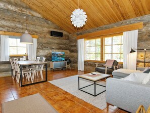 Interior rústico de una cabaña con comedor, sala de estar con sofá y sillones, y un amplio ventanal con cortinas.