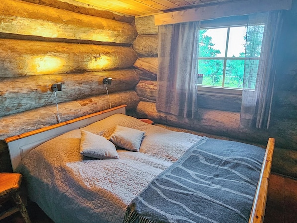 Une chambre dans un chalet en rondins, avec un lit, des oreillers et une fenêtre avec des rideaux.