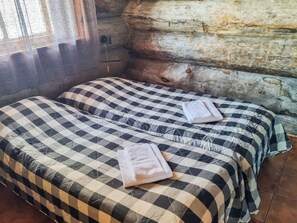 Une chambre avec deux lits, chacun recouvert d’une courtepointe à carreaux et agrémenté d’une serviette pliée. La chambre est dotée d’un mur en bois brut et d’une fenêtre avec un rideau.