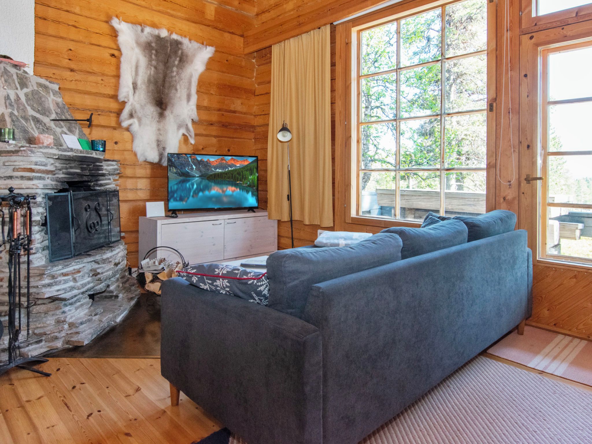 L’intérieur chaleureux d’un chalet avec une cheminée en pierre, un téléviseur et une grande fenêtre donnant sur le paysage extérieur.