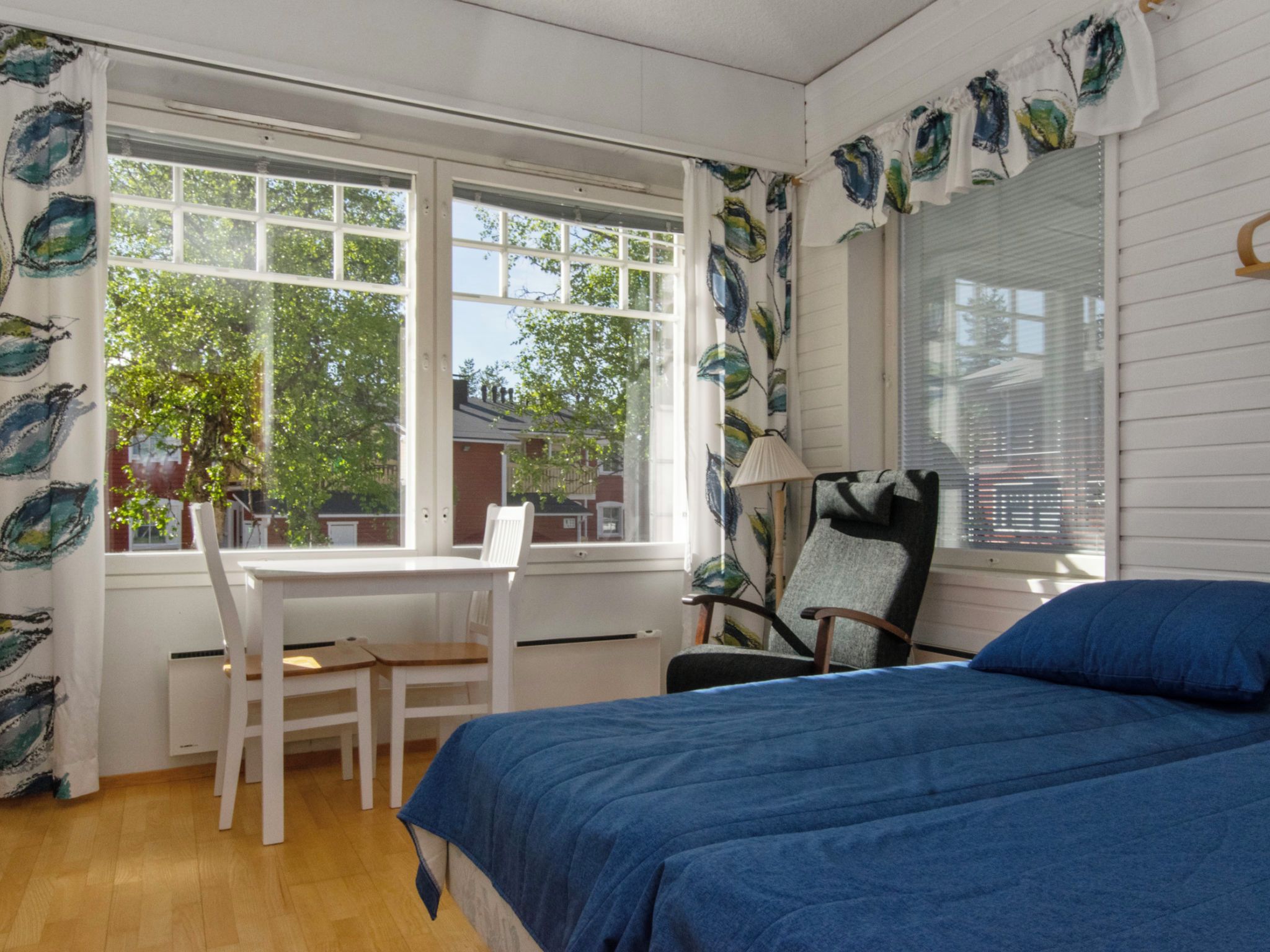 A room with a bed, a desk, and a chair. There are windows with curtains.