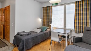 Ein kleiner, hell erleuchteter Raum mit zwei Betten, einem Schreibtisch und einem Stuhl. Ein Fenster mit Karottenvorhängen und ein darüber befindliches Schild mit der Aufschrift 