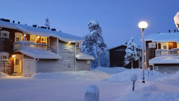 A snow-covered residential area with houses, a street lamp, and a mailbox.