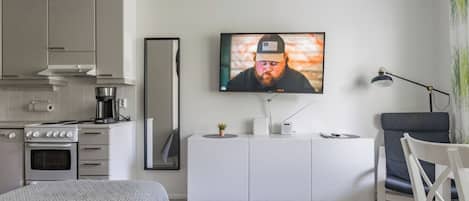 Una sala moderna con un televisor de pantalla plana montado en la pared, un comedor con mesa y sillas, y una kitchenette con estufa y horno.