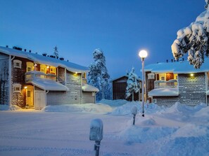 Lumipeitteinen mökkikompleksi, jossa on useita rakennuksia, hyvin valaistu sisäänkäynti ja lumipeitteinen maisema.