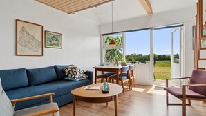 Ein modernes Wohnzimmer mit einem blauen Sofa, einem Holztisch und einem Essbereich mit Tisch und Stühlen. Große Fenster bieten Blick ins Freie.