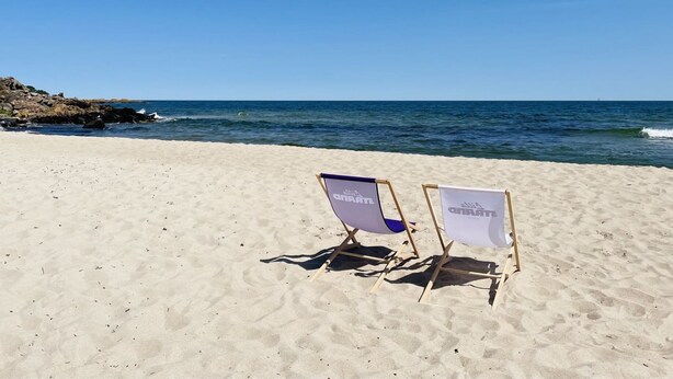 Zwei Strandstühle auf sandigem Strand mit felsiger Küstenlinie und ruhigen Meereswellen im Hintergrund.