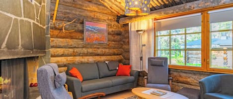 A log cabin interior with a stone fireplace, a sofa, a chair, a table, and a window with a view of greenery.
