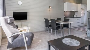 A modern living room with a flat-screen TV, a grey armchair, a dining table with chairs, and a kitchen area with cabinets and a stove.