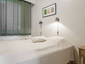 Een net opgemaakt bed met een witte deken, een kussen en een ingelijst beeld erboven. Er is een raam met een groene gordijn en een wandlamp.