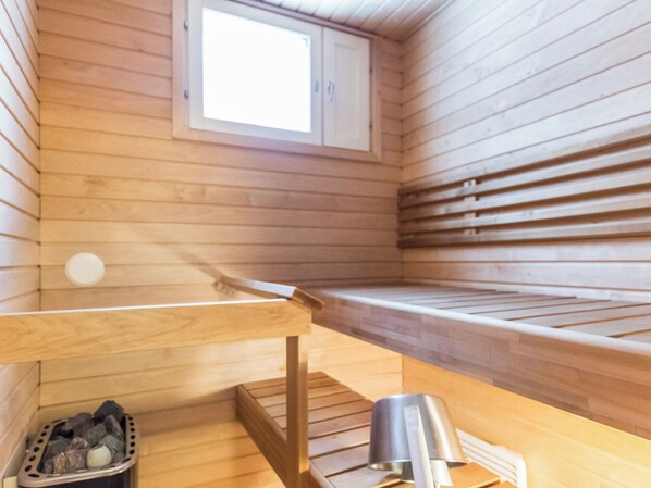 A sauna room with wooden walls, a bench, a small window, and a metal container on the floor.