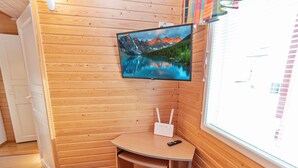 A cozy room with wooden walls, a mounted TV, a small desk with a chair, and a window with blinds.