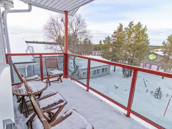 A covered outdoor area with a snow-covered deck, wooden chairs, and a view of a snowy landscape with trees and buildings. - Apartment helmi saariselkä by Interhome (Inari)