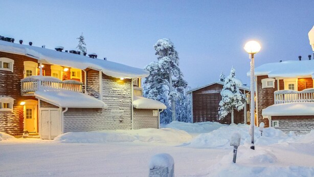 A snow-covered residential area with houses, a wooden barn, and a street lamp.