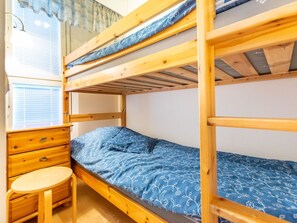 A bunk bed room with a wooden desk and chair, a window with blinds, and a patterned bedspread.