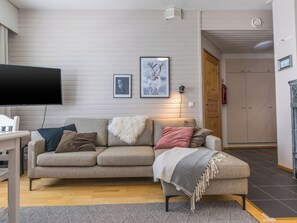 A living room with a sofa, ottoman, and a television. There are framed pictures on the wall, a lamp, and a door leading to another room.