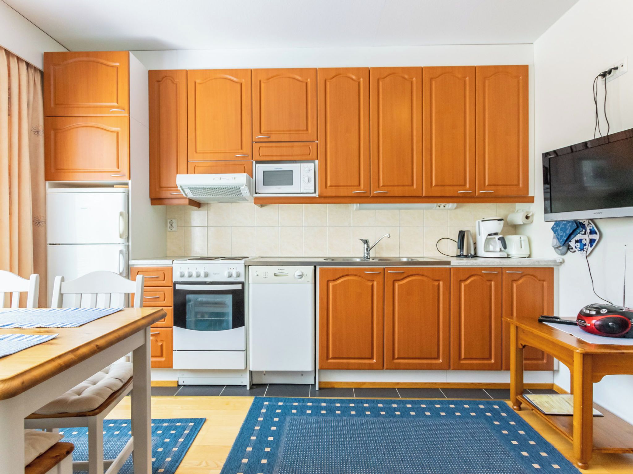 Cocina compacta con armarios de madera, microondas, estufa, lavavajillas y televisor en la pared.