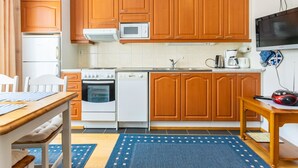 A compact kitchen with wooden cabinets, a microwave, a stove, an oven, and a dishwasher. There is a dining table with chairs and a blue rug on the floor.